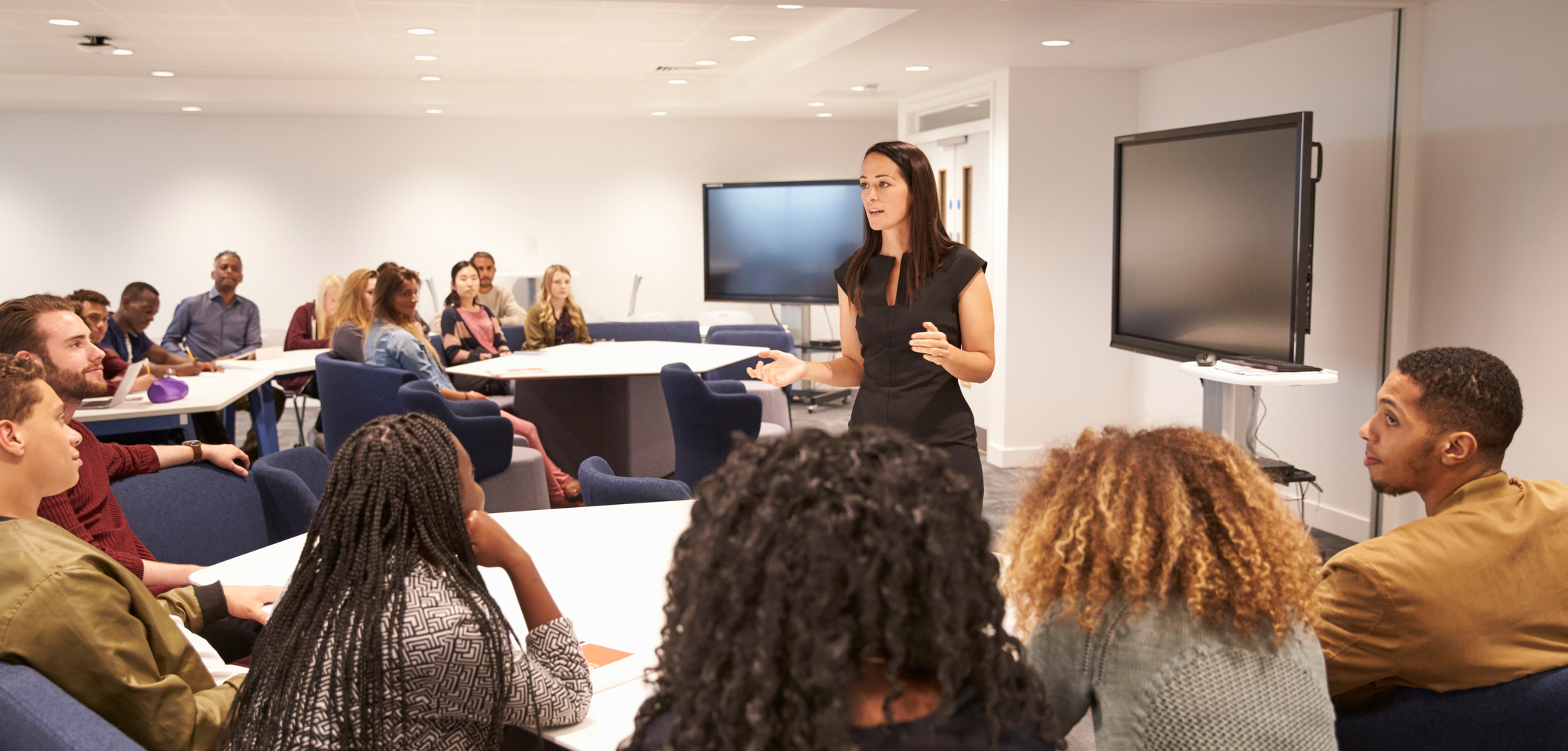 woman wearing a black dress speaking in front of a group of people in a classroom setting