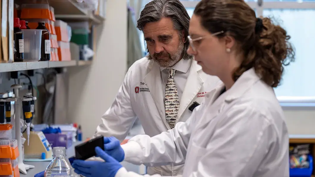 a clinical researcher watches as another researcher performs lab tests