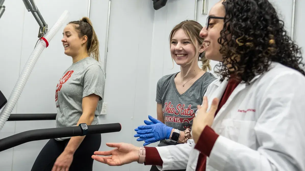 female undergrad researcher in white lab coat testing two young women on a treadmill