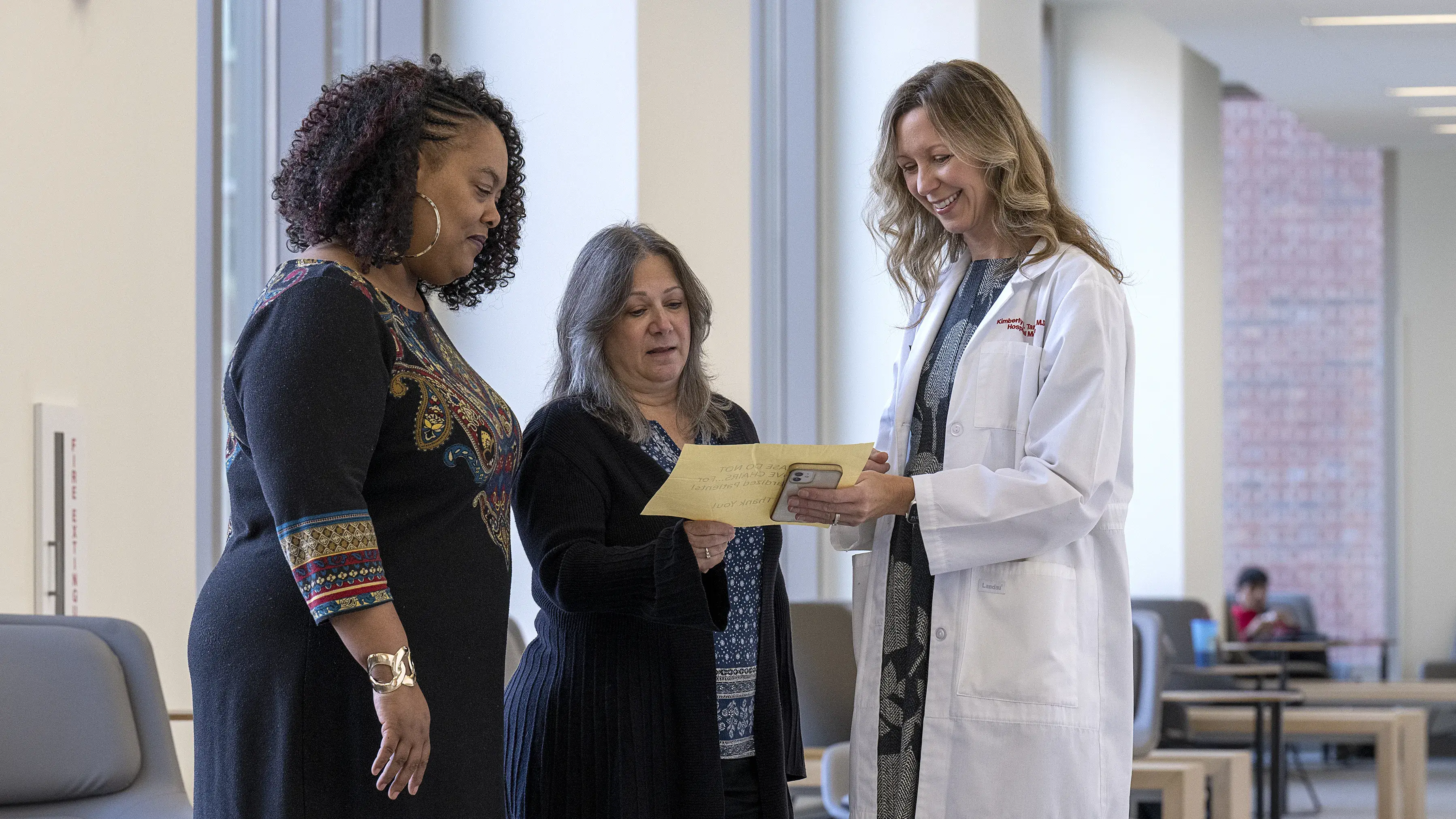 Woman in lab coat speaking with two women in business casual clothes in a modern lobby